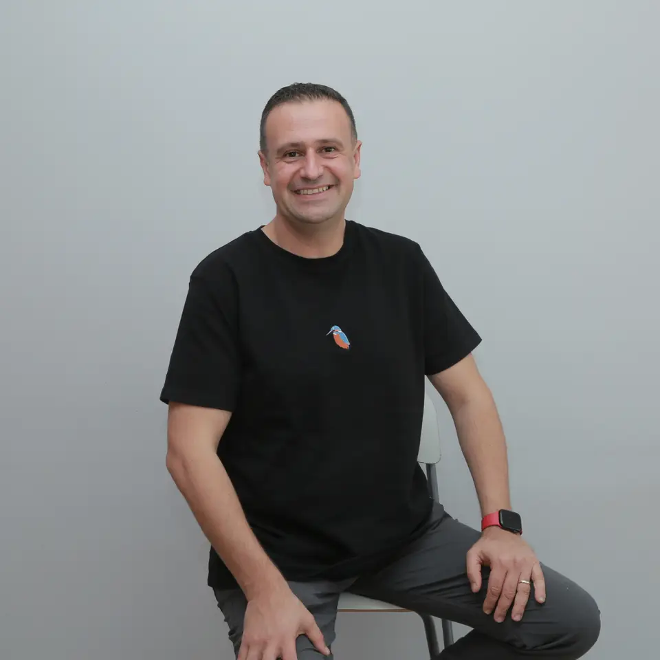 Neşet Birlik in a black T-shirt, sitting on a chair and smiling
